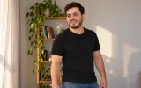 A young man in a plain black t-shirt in a bright apartment